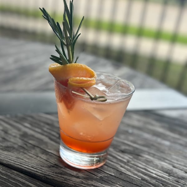 A pink cocktail with ice, garnished with orange peel and a sprig of rosemary on a wooden table.